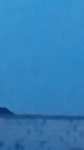 This media file shows a blurry, low-light outdoor scene. A dark, triangular object, possibly a building or a mound, is visible on the left side of the frame, silhouetted against a muted, overcast sky. The ground in the foreground is indistinct, appearing as a hazy expanse of white and blue, suggesting a flat, open area. There are no discernible people or clear objects in the scene. The overall impression is one of a desolate landscape under a dim sky. The time of day appears to be either dawn or dusk, or possibly a very cloudy day. The location context provided is "Іргелі ауылдық округі, Kazakhstan." No text is visible in the media file.