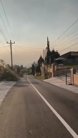A vertical video shows a paved road curving uphill with a white line down the center. Power lines crisscross the sky. On the right side of the road, a stone wall topped with red brick pillars and fencing runs alongside the road, with vegetation growing along the base and over the wall. Beyond the wall, there is a courtyard with paved walkways, outdoor seating with umbrellas, and a building in the distance. Tall cypress trees are planted along the top of the wall. On the left side of the road, there are trees and a power pole. The sky is clear with a pale orange hue, suggesting either sunrise or sunset. The road appears to be in a rural or suburban mountainous area.