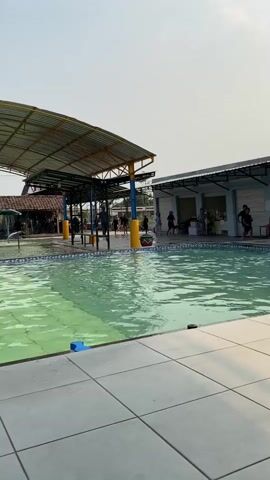 The video shows an outdoor swimming pool area. The pool water is green. There is a tiled area around the pool. To the left, there is a covered structure with a yellow roof supported by blue and yellow poles. Several people are visible under this structure and near the pool's edge. To the right, there is a building with a gray roof and open-sided sections. People are gathered under this structure as well. In the background, there is a clear sky and some trees. The scene appears to be a public pool or water park on a bright day.