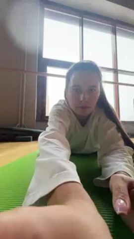 The video shows a woman on a green yoga mat in what appears to be a dance or exercise studio. She is wearing a white long-sleeved shirt and her hair is partially pulled back. She leans forward from a prone position, placing her head on the mat. There's a barre along a wall with a large window in the background. The lighting is bright, likely from the window.
