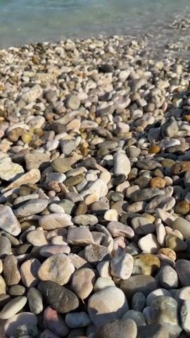 The video shows a pebble beach with gentle waves lapping at the shore. In the background, several people are swimming and enjoying the turquoise water. Hills can be seen in the distance under a clear blue sky. The camera is positioned low to the ground, focusing on the smooth, varied stones that make up the beach.