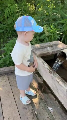 A toddler wearing a blue cap and a light-colored t-shirt stands on a wooden platform next to a trough of water. The toddler is reaching towards the water with their right hand. Water is flowing into the trough from a pipe. Green foliage is visible in the background.