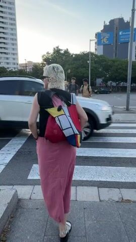 The video captures an urban street scene in L'hospitalet De Llobregat, Spain, during late afternoon or early evening. A woman with short blonde hair and sunglasses, dressed in a black top, pink maxi skirt, and a distinctive geometric-patterned backpack (resembling a Mondrian painting), is seen crossing a zebra pedestrian crosswalk. Two young men follow her. A white car passes by on the right.

Tall buildings dominate the background, including a modern structure with black and reflective glass panels that displays "COPISA" near its top. Graffiti, including "Snap," "Umbro," and "TAFE," is visible on concrete walls and barriers along the street. A blue billboard with text like "bluepay" and "1€" is also visible in the distance. The camera pans to show the broader cityscape, revealing more high-rise buildings and road infrastructure, including clear white road markings on the asphalt. The sky is clear and light blue, with soft sunlight illuminating parts of the buildings.