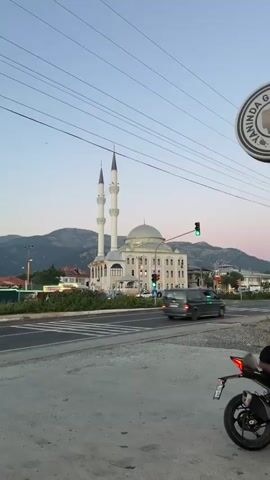 The video shows a mosque with two minarets and a dome, standing across a busy street with traffic signals. The mosque is predominantly white or light beige with a grey dome. Mountains form a backdrop. A motorcycle is partially visible in the foreground. Power lines crisscross the sky. A car and a van drive across the intersection. The sky is light, indicating early morning or late evening. The adhan, the Islamic call to prayer, can be heard.