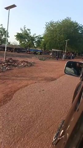 The video shows a scene in a rural or semi-rural area. The road is primarily red dirt, with some segments of paved road. Several people are walking and riding motorcycles in the vicinity. There are several structures, including simple buildings and sheds, some surrounded by stacks of tires and miscellaneous items. A few larger trees are present, and the sky is clear. The video is taken from inside a moving vehicle, showing the driver's side mirror.