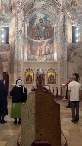 In a dimly lit interior of what appears to be an ancient church, several individuals are gathered. The scene is dominated by ornate frescoes covering the arched walls and ceiling, depicting religious figures and scenes in muted colors. In the foreground, a large, intricately carved wooden object, possibly a choir stall or a lectern, faces the altar.

The altar area itself is adorned with icons, candles, and a central religious artifact. A few people, some with their backs to the camera, are observing the altar, suggesting a moment of prayer or contemplation. One woman is wearing a headscarf and a green skirt, while another wears a dark headscarf and coat. A man in a white t-shirt stands to the right, facing the altar.

The lighting is low, with artificial lights illuminating sections of the frescoes and the altar area. The overall atmosphere is one of solemnity and reverence, indicative of a religious service or a visit to a sacred site. The location cues, such as the specific style of frescoes and iconography, along with the provided context of Nikortsminda, Georgia, point to this being a historical Georgian Orthodox church. There is no visible text within the frame.