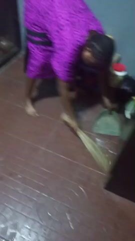 A person wearing a purple dress sweeps the floor with a broom. The person is bent over, actively sweeping in the lower right corner of the frame. In the background, there is a counter with various kitchen items, including pots and containers. The floor is tiled.