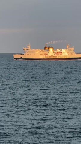 A wide shot captures a large white ferry boat sailing across the deep blue ocean. The ferry is identified with the name "Scandlines HYBRID FERRY" in red and black lettering along its side. Plumes of white smoke or steam billow from the ferry's smokestack, drifting towards the right side of the frame. The ferry is illuminated by the warm glow of the setting sun, which casts a golden hue across its hull and the water. The sky above is a hazy grey with hints of pink and orange, suggesting the sun is low on the horizon. The water is choppy with visible waves.