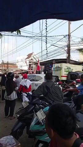 The video shows a street scene with several people on motorcycles and some standing. There are Indonesian flags displayed on poles, and a large speaker system is visible on a truck in the background. The atmosphere appears to be lively, possibly indicating a public event or celebration. The video is shot from a slightly elevated, shaky perspective, suggesting it was recorded on a mobile device while in motion. The environment is characterized by overhead wires and buildings, typical of an urban or semi-urban setting.