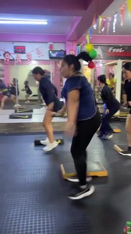 A group of women, mostly wearing athletic attire, are participating in a fitness class within a gym. The room has pink walls and a mirrored wall reflecting the participants and the space. Several women are positioned on yellow aerobic steps, performing exercises that involve squatting, jumping, and arm movements. One woman in the foreground is wearing a blue top and black leggings and is holding a set of small, colorful balls. Another woman in a black top and shorts is also on a step. In the background, more women are engaged in similar exercises, and there is gym equipment visible, including a treadmill. A digital clock displays the time, and colorful banners hang along the top of the room. The floor is dark with a textured pattern. The overall atmosphere suggests a lively and energetic workout session.