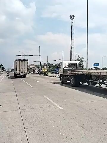 The video shows a road scene at an intersection, possibly in a city. Several vehicles are present including large trucks and motorcycles. Traffic lights are visible and appear to be red. A person on a motorcycle is shown riding through a crosswalk. The sky is partly cloudy. There are signs and roadside vegetation at the intersection. The overall impression is of an average urban scene with moderate traffic.