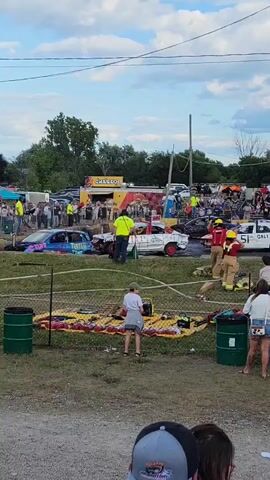 A demolition derby event is underway. Several cars are present in the arena, some of which appear damaged. A group of individuals wearing yellow vests and helmets are present, some carrying equipment, possibly safety personnel. Another group, dressed in red and yellow firefighting gear, are also in the arena, seemingly attending to a car. Spectators are visible in the background, with some standing behind a fence. There are also food vendors and tents set up. The sky is cloudy.