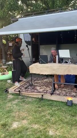 A group of people are gathered outdoors, possibly at a festival or outdoor event. In the foreground, on a raised wooden platform covered with rugs, two individuals are operating DJ equipment. One person, wearing a grey top and tan pants, stands behind a laptop. To their right, another person, in a black t-shirt, is actively engaged with DJ turntables. Behind them, to the left of the platform, a person wearing a dark outfit and an orange hat is holding a microphone and appears to be singing or speaking into it. The background shows a structure that resembles a small building or food truck with open sides, decorated with string lights. The setting is grassy with trees visible in the distance, suggesting an outdoor, possibly casual, gathering.