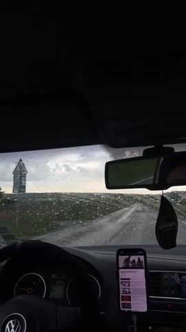 Here's a description of the video:

The video shows a view from inside a car driving on a wet road. It appears to be raining, as there are raindrops on the windshield. The windshield wipers are activated. In the center of the dashboard, a smartphone is mounted, displaying a music playlist.  A black air freshener is hanging from the rearview mirror. In the reflection of the rearview mirror, a person's eyes are visible, likely the driver. The dashboard and steering wheel of the car are visible in the foreground. The external environment is blurry with dark skies and some visible green fields adjacent to the road. The interior of the car is dark, contrasting with the brighter scene outside.