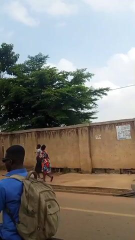 The video shows a moving perspective, likely from a vehicle, along a street with a concrete wall on the right. Lush green trees with full canopies are visible behind the wall. Several people are seen walking or standing along the sidewalk. One individual, wearing a red outfit, stands near the wall. Further down, another person in dark clothing walks away from the camera. The wall itself appears aged and weathered, with some sections showing signs of disrepair and exposed brickwork. A tall, thin wooden pole stands next to the wall, with a wire extending horizontally from its top. The sky is partly cloudy. The overall scene suggests an outdoor, urban or semi-urban environment.