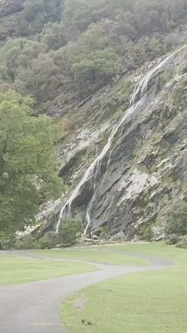 A very tall, thin waterfall cascades down a rocky, moss-covered mountainside. Dense green foliage covers the top and sides of the mountain. The base of the waterfall leads to a grassy area with a paved path winding through it.