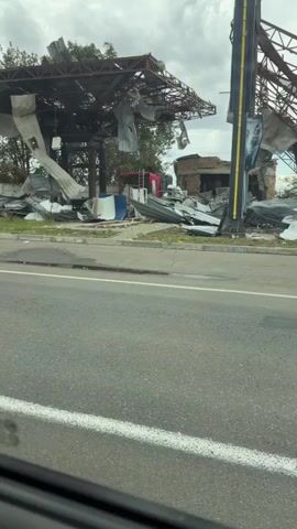 The video shows a damaged structure, likely a gas station, with significant debris and structural damage. The roof is largely collapsed and torn, with large pieces of metal hanging precariously. Building walls are broken and incomplete, and rubble is scattered across the area. A red fire truck is visible in the background, partially obscured by debris. The overall environment appears to be overcast and possibly windy, with trees visible in the background also showing signs of damage. The perspective suggests the footage is being filmed from inside a vehicle moving past the scene.