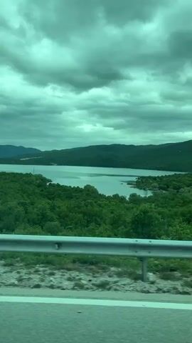 The video depicts a view of a lake surrounded by dense green forests, taken from a moving vehicle. The sky is overcast with thick, gray clouds. A guardrail is visible in the foreground. The lake reflects the cloudy sky. The footage is slightly tinted in green.
