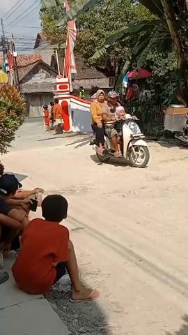 The video shows a street scene with several people. In the foreground, two young boys are sitting on the ground, one wearing an orange shirt and black shorts, the other wearing a black hat and a black shirt. They appear to be watching something happening further down the street. In the background, a motorcycle carrying three people, an adult male driver, an adult female passenger, and a child passenger, is moving away from the camera. To the left of the street, there is a white structure with red and white striped decorations and what appears to be a finish line banner. Several other people, including women and children, are standing near this structure, some of whom are dressed in traditional clothing and headscarves. The environment is a residential street with houses and trees visible. The ground is a dirt or gravel road. The overall atmosphere appears to be festive or celebratory.