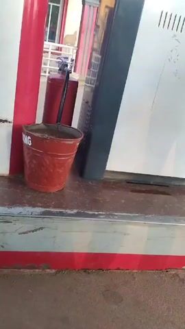 The camera pans up along a gas pump at a gas station. The lower part of the pump is red and white. There is a red bucket that says “Smoking” on it beside the pump. The pump itself is white and grey. A woman wearing a grey t-shirt and black leggings walks behind the pump. She is holding a gas hose.