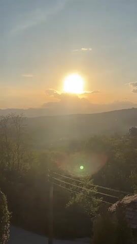 The video shows a landscape during a sunset. The bright sun is partially obscured by clouds near the horizon. Rolling hills are visible in the distance, and the foreground is filled with lush trees and vegetation. A small, old building with a red tile roof sits amidst the greenery. The camera pans slowly across the scene, revealing more of the landscape and the building. A tall, slender tree stands out among the other trees. As the camera continues to pan, a white structure with an arched opening comes into view in the lower left corner of the frame.