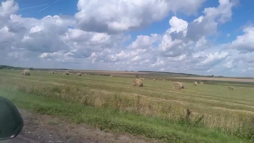 The video shows a view from a vehicle driving along a dirt road. The road is on the left side of the frame and is partially visible. To the right of the road is a field of green grass with several round hay bales scattered across it. The field extends into the distance, meeting a gently rolling landscape under a blue sky with fluffy white clouds. There are no people or animals visible in the frame. The sky has some faint white contrails from airplanes. The overall impression is a rural, agricultural landscape on a bright, partly cloudy day.