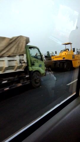 The video shows a close-up shot of construction equipment on a road. In the foreground, a light green flatbed truck is visible, carrying a large, covered load. The truck is moving, with its wheels in motion. Behind the truck, a yellow road roller with the brand name "SAKAI" is present, appearing to be stationary. Further behind the road roller, another piece of construction equipment, possibly a smaller roller or a similar vehicle, is visible. The road surface appears to be asphalt. The scene is viewed from a moving vehicle, indicated by the slight blur and the reflection in what seems to be a window. The overall environment suggests an outdoor construction or road maintenance site.