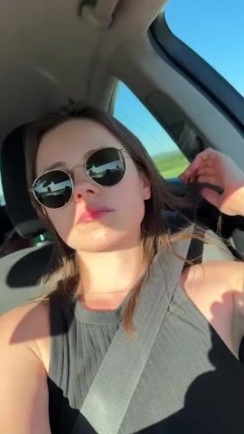 Here is a description of the video:
A woman wearing a black tank top and round sunglasses is sitting in the driver's seat of a car. She has her seatbelt on. She touches her hair, poses for the camera by putting her hand behind her head and puckering her lips, and then waves at the camera. The sky is visible through the car window.