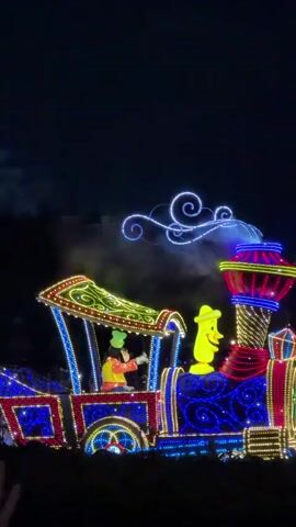 A nighttime parade float, elaborately decorated with bright, multicolored lights, moves across a dark outdoor space. The float resembles a train with a locomotive and several passenger cars. A character, possibly Goofy, wearing a hat and orange shirt, is visible in the locomotive's driver's seat. Another character, dressed in red and white polka dots with a bow in her hair, resembling Minnie Mouse, stands on a platform on a subsequent car. Other figures can be seen on the float's lower levels. Many people are gathered in the foreground, watching the parade, with some holding up cell phones to record the event. In the background, distant structures are visible against the dark sky, and wisps of white smoke or fog are present. The overall atmosphere is festive and celebratory.