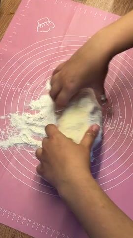 The video shows a close-up, overhead view of a pair of hands kneading dough on a pink, circular baking mat with measurement markings. The dough is partially covered in white flour. The hands are actively working the dough, pushing, folding, and manipulating it. The environment appears to be a kitchen or food preparation area, with a wooden surface visible beneath the pink mat. The lighting is bright, highlighting the flour and the texture of the dough.