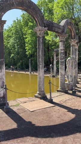 The video captures a serene and historically rich scene under a bright, sunny sky. The camera pans from left to right, revealing a long, rectangular pool of brownish-yellow water, flanked by lush green trees. In the foreground, weathered stone columns and arches, characteristic of ancient Roman architecture, frame the view.

As the camera pans, a prominent white marble statue comes into full view, standing between two large, ornate columns. The statue depicts a male figure, partially nude, wearing a helmet and holding a shield, with its back to the viewer. This artistic element adds to the classical ambiance of the setting.

Along the far bank of the pool, several small figures of people can be seen walking, enjoying the historical site. At the very end of the long pool, a small, domed structure is visible amidst the trees, likely a nymphaeum or grotto. The overall impression is one of a grand ancient villa or garden. The chains between the columns in the foreground suggest an area where access to the immediate edge of the pool might be restricted.

The clear blue sky and bright sunlight indicate that it is daytime, and the weather is pleasant. This location, with its distinctive columns, statues, and reflecting pool, is unmistakably the Canopus at Hadrian's Villa in Tivoli, Italy. No visible text appears in the video.