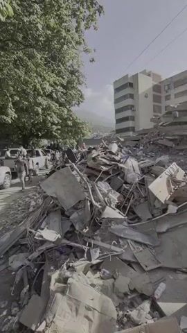 The video pans across a scene of destruction, with large piles of rubble and debris dominating the foreground. In the background, a multi-story building stands partially damaged, with several floors collapsed. A few people are visible amongst the rubble, some appearing to be rescue workers or survivors. A white appliance, possibly a washing machine, lies on its side near the debris. An excavator is also visible in the distance, suggesting ongoing cleanup or rescue efforts. The sky is clear and bright, with the sun visible, casting long shadows. The overall impression is one of devastation and the aftermath of a significant event, likely an earthquake or other disaster.