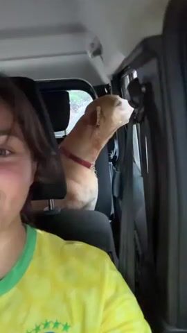In this short video, a young woman is in the front seat of a car taking a selfie. She is wearing a yellow and green floral patterned shirt. In the back seat is a medium sized, light brown dog with a red collar. The dog is sitting upright and looking out the window, panting. The woman looks at the dog and smiles. She reaches back and pets the dog on the head.