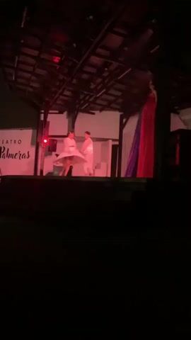 This is a video clip of a performance on a stage. Two dancers in white costumes are on stage. The stage has a wooden roof structure and a white backdrop with the words "TEATRO" and "las Palmeras" visible. There are red and blue drapes on the right side of the stage. The lighting is predominantly red and green. The dancers are interacting with each other as if performing a routine.