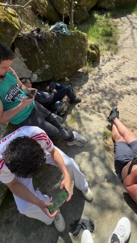 The video shows a group of men outdoors, possibly in a park or wooded area, relaxing and using their phones. In the first scene, two men are sitting on the ground looking at their phones while another lies down in the background. Then, the camera shifts to a man lying on the ground wearing an aluminum foil hat, also looking at his phone. Two other men are standing nearby. An empty bottle lies on the ground. The overall atmosphere appears casual and relaxed.