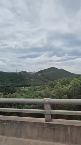This is a video taken from a moving vehicle, looking out over a landscape. In the foreground is a concrete barrier with metal railings. Beyond that, a verdant valley is visible, filled with trees. In the distance, rolling green hills rise up, partially obscured by a cloudy sky. A few power lines are visible on the left side of the frame, and a large construction crane appears on the right side for a brief moment. The overall impression is one of a natural, possibly rural, setting under an overcast sky.