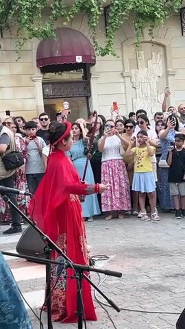 A woman in a red traditional dress plays a stringed instrument in front of a crowd of people. The woman is wearing a red headscarf and her hair is pulled back in a bun. She is holding the instrument with her right hand and bowing it with her left. The crowd is made up of men, women, and children of various ages. Most of the people in the crowd are holding up their phones to take pictures or videos of the performance. The scene appears to be taking place on a street, with a building in the background. There are plants hanging from the building and a red awning over the entrance. There is also a microphone stand in the foreground.