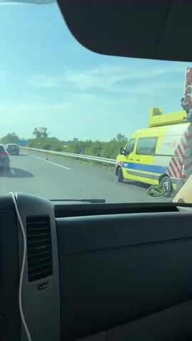 The video is filmed from the interior of a vehicle, looking out through the windshield. The vehicle is stationary in a traffic jam on a highway. In front of the vehicle, there are several other cars. To the right, there is a yellow and blue van that appears to be an emergency vehicle, possibly an ambulance or police van, with its lights flashing. The road is lined with trees and vegetation. The sky is clear and blue, indicating a sunny day. The traffic appears to be moving slowly or not at all. Orange traffic cones are visible on the side of the road.