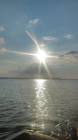 The video shows a view from the back of a boat sailing on a body of water. The sun is bright and shining, casting a reflection on the water. The sky is blue with some scattered clouds. The horizon is visible in the distance, with land appearing as a faint line. A person's legs are visible in the foreground, indicating that they are sitting in the boat. The boat appears to be moving, as evidenced by the slight wake behind it.