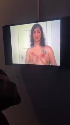 A person is viewing a video on a flat screen television. The video shows a topless person with dark hair, light skin, and dark eyebrows, who is looking directly at the camera. The person has tattoos on their arms and upper chest, and there appear to be red marks on their chest. The background appears to be a room with a closed white door. The person viewing the television is silhouetted and appears to be wearing a dark garment. The room is dimly lit.