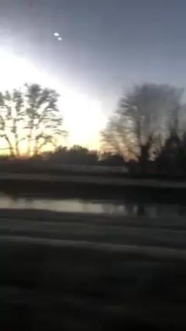 The video shows a landscape passing by, seen from a moving vehicle. Silhouetted trees are visible against a light-colored sky, which transitions from gray to a brighter hue near the horizon, possibly indicating sunrise or sunset. There appears to be a body of water or a reflective surface in the foreground. The ground and road are dark and blurry due to the motion. There are no people visible in the video.