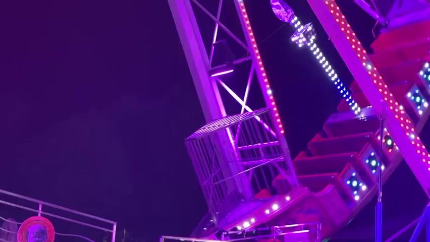 This media file captures a thrilling amusement park ride at night, likely in Riccione, Italy. The ride, a large pendulum attraction resembling a ship or a Viking vessel, is adorned with a multitude of bright, flashing lights in red, green, and blue. The structure itself is a complex metal framework, painted in vibrant colors, with the illuminated ride vehicle visible at the top of its arc.

The scene is set against a dark night sky, indicating the event is taking place after sunset. There is no visible indication of weather conditions, but the darkness suggests a clear evening. The ground appears to be asphalt, with some sections illuminated by the ride's colorful lights.

While there are no clearly identifiable people within the ride's passenger cars, a figure is visible in a caged area on the main structure, possibly an operator or a park attendee. The overall impression is one of excitement and exhilaration, typical of a night at an amusement park. The bright, dynamic lighting and the visible motion of the ride contribute to a lively and energetic atmosphere. There is no visible text in the media file.
