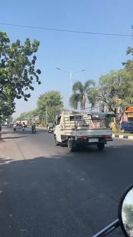 A white pickup truck is driving down a paved road on a sunny day. The truck is carrying a long, flat object that resembles a ladder or metal sheeting on its roof rack. The truck's bed is empty. Behind the truck, a motorcycle with a yellow helmet is also traveling. The motorcycle has two large canvas bags attached to the rear. A few trees are visible on the side of the road, and streetlights are present. The road has lane markings.