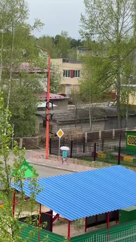 The video captures a medium shot from an elevated perspective, likely from a building overlooking a neighborhood scene on an overcast day. A person with a light blue umbrella is walking on a sidewalk, passing by a yellow diamond-shaped warning sign and a red and orange utility pole with electrical equipment attached. The neighborhood consists of low-rise buildings, some with red roofs and yellow exteriors, nestled among green trees, typical of a residential area. In the foreground, a playground structure with a blue corrugated metal roof and green fencing adds a touch of vibrancy to the scene. Overall, the setting seems peaceful and typical of a suburban or small-town environment.