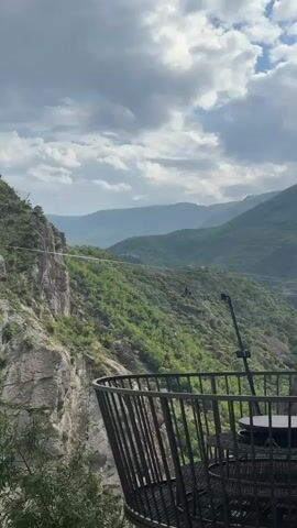 A vertical video shows a mountain range with lush green trees and a winding river at the base of a cliff. A long white cable stretches across the valley, connecting two mountains. People are visible in the distance, standing on a platform or structure on the opposite mountain, possibly preparing for an activity like zip-lining. In the foreground, a dark metal railing of a viewing platform or balcony extends into the frame, with a small table and a dark object (possibly a camera or tripod) visible behind it. The sky is partially cloudy.