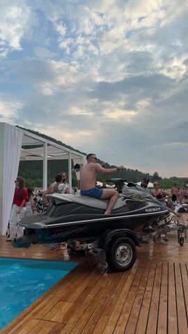 The video shows a man sitting on a jet ski that is on a trailer. He is shirtless and wearing blue swim trunks. He is pointing his right arm towards the right side of the frame and looking in the same direction. The jet ski is black and gray. It is parked on a wooden deck next to a pool. In the background, there are several people standing around, some of whom are looking at the man on the jet ski. There is also a white tent structure in the background. The sky is cloudy and appears to be late in the day.
