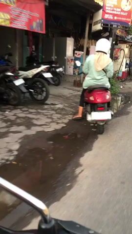 The video shows a person riding a red scooter down a street in Balonggabus, Indonesia. The person is wearing a white helmet and a light green shirt with a tan headscarf. The scooter has a white license plate with blurry text.

The scene is set along a street with shops on the left. Several other motorcycles are parked in front of the shops. The ground is wet, suggesting recent rain. The time of day appears to be late afternoon or early evening, as indicated by the lighting.

One of the shops has a red banner with text that is difficult to read but appears to be in Indonesian. Another sign above a shop reads "BUKU YASIN UNDANGAN KILAT". The overall atmosphere is one of everyday life in a bustling street market or commercial area.