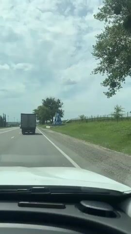 The video captures a roadside view from a moving vehicle. A large sign with the inscription "АНАПА" (Anapa) and "ДОБРО ПОЖАЛОВАТЬ" (Welcome) is prominently displayed on the right side of the road. The sign is stylized with blue and white geometric patterns and resembles a sailboat. To the left of the sign, a large tree with sparse leaves stands. Beyond the sign, a grassy slope extends, bordered by a fence. The road itself is paved with white lane markings. A white vehicle is partially visible in the foreground, indicating the perspective is from inside a car. The sky is overcast with clouds. A truck is seen in the distance on the left side of the road. The overall scene depicts an entrance to a town or city.