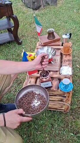 The video shows a person outdoors on grass, preparing to grind coffee beans. They are holding a metal bowl full of roasted coffee beans in their left hand, and with their right hand, they are adding beans to a manual coffee grinder placed on a wooden pallet. To the right of the grinder, there's a small stove with a metal coffee pot on top. Further to the right, there are two coffee cups and saucers, one empty and one with a blue design. There are also two small flags, one of Iran and one of Moldova, on sticks placed behind the coffee-making setup. A child is standing to the left of the person, observing the process. The background shows more grass and a part of a wooden structure.