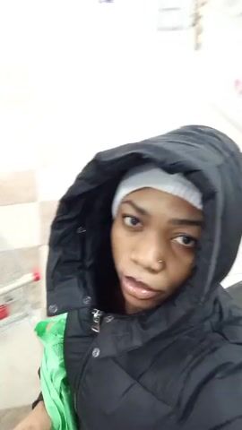 The video shows a person wearing a dark puffer jacket with the hood up. Underneath the hood, they are wearing a white beanie. The person is looking directly at the camera.  The background changes slightly, showing hints of a store interior with overhead lights and a checkout area in some frames. The camera seems to be held by the person, as there is slight movement.